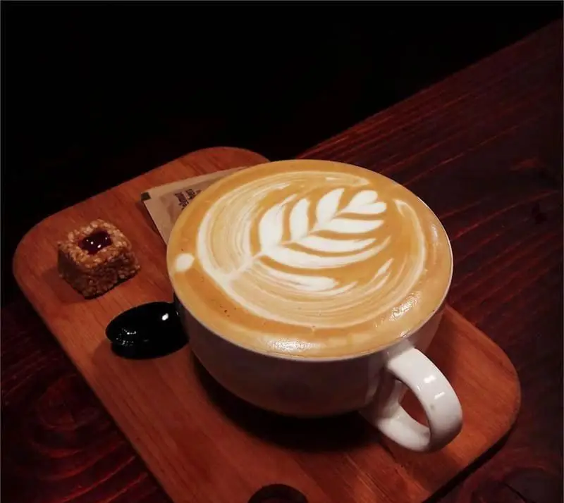 قهوه لاته خوش‌طعم با هنر لاته چشم‌نواز (طرح برگ) روی تخته چوبی در کافه چری. همراه با یک شیرینی کوچک و قاشق برای لحظاتی آرامش‌بخش.