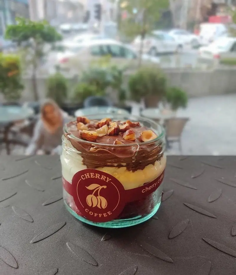 دسر لایه‌ای شکلاتی و خامه ای خوشمزه کافه چری با روکش فندق، داخل ظرف شیشه‌ای، مناسب برای یک میان وعده دلپذیر.