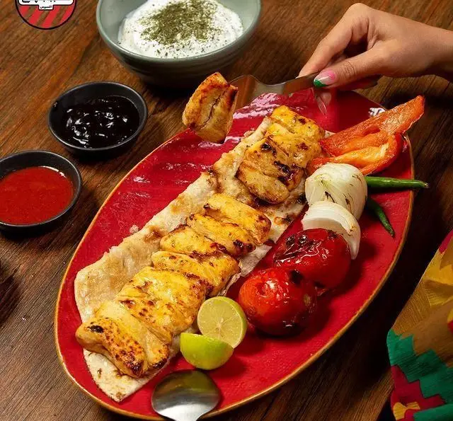 جوجه کباب خوشمزه کبابچی آجودانیه با نان، گوجه، پیاز کبابی، لیمو، سس‌های متنوع و ماست در یک بشقاب قرمز چوبی. تجربه‌ای لذیذ از غذای ایرانی.