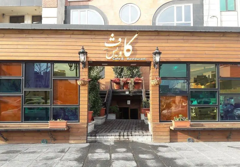 نمای بیرونی کافه رستوران کاویش با دکور چوبی، پنجره‌های رنگی جذاب و ورودی مزین به گلدان و فانوس‌ها.