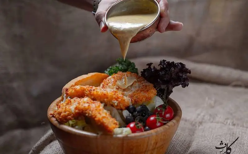 تصویری از سرو سالاد مرغ سوخاری کافه رستوران کاویش با سس مخصوص، گوجه گیلاسی، زیتون و نان. غذایی لذیذ و تازه.
