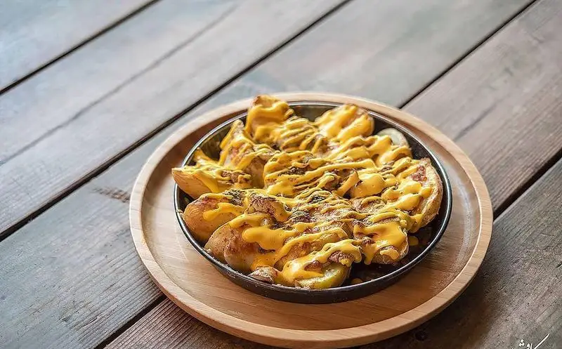 سیب زمینی پنیری مخصوص کافه رستوران کاویش، همراه با سس پنیر آب شده طلایی و سس مخصوص، سرو در تابه روی سینی چوبی.