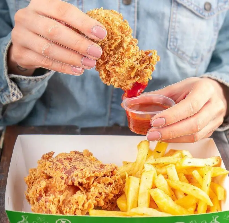 لذت مرغ سوخاری طلایی چیکولند: دستانی در حال دیپ کردن تکه‌ای ترد در سس قرمز، در کنار سیب زمینی و تکه دیگر مرغ سوخاری.