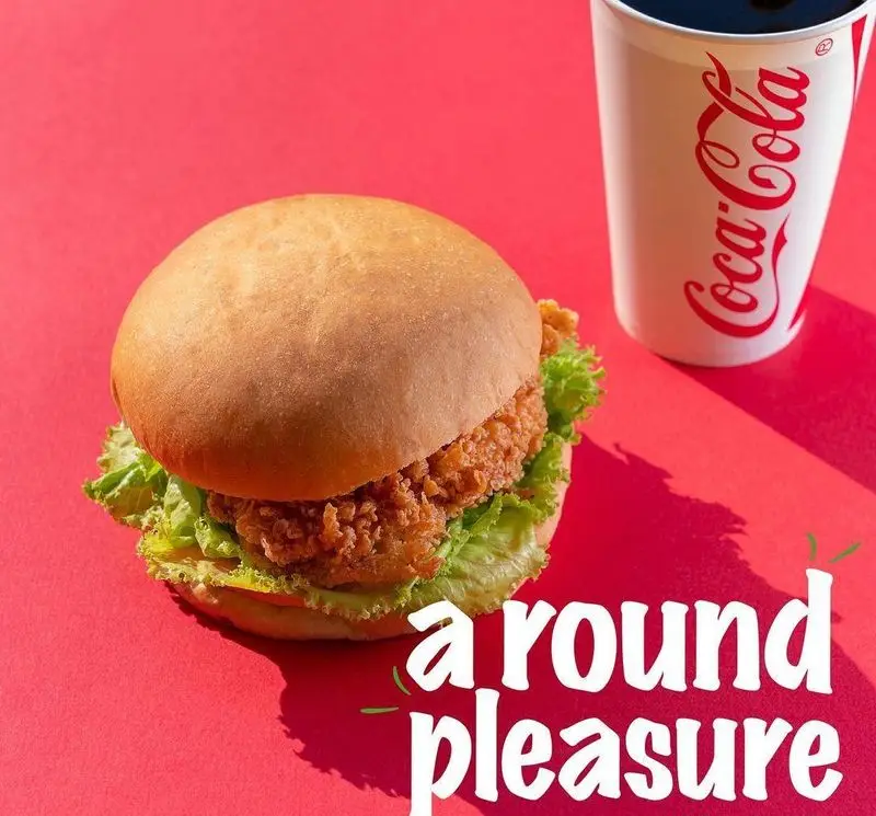 همبرگر مرغ سوخاری تازه و خوشمزه از فست فود چیکولند، در کنار نوشابه کوکاکولا روی پس‌زمینه قرمز. لذت یک وعده عالی.