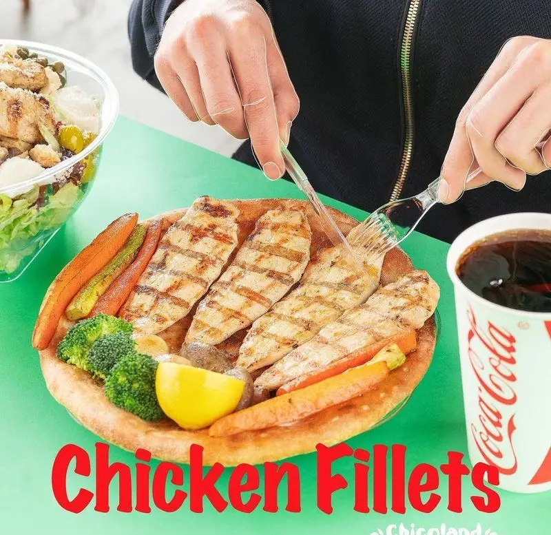 فردی در حال برش فیله مرغ گریل چیکولند، سینه مرغ تازه با سبزیجات کبابی، سالاد و نوشابه. وعده‌ای سالم از فست فود چیکولند.