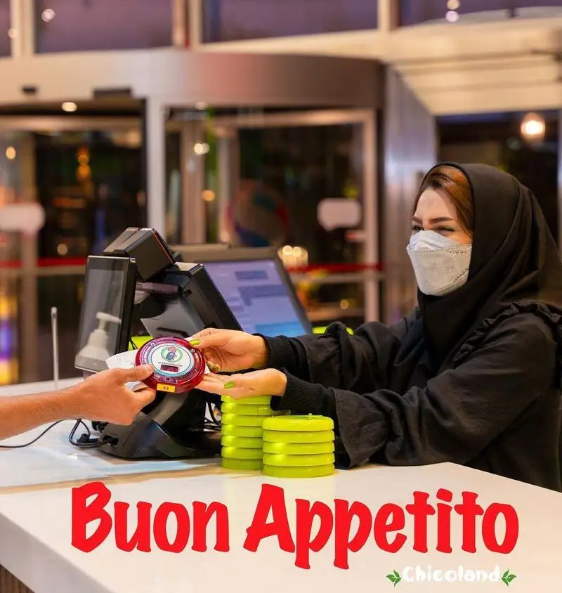 کارمند ماسک‌زده فست فود چیکولند در حال تحویل پیجر سفارش به مشتری در پیشخوان، با نمایش پیجرهای سبز و دستگاه پوز.