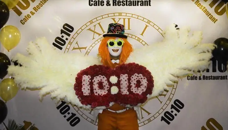 دکوراسیون خاص رستوران فرنگی ده و ده با تم ساعت ۱۰:۱۰، شامل فردی با لباس مبدل بالدار و گل‌آرایی 10:10 در پس‌زمینه ساعت بزرگ و بالن‌ها.