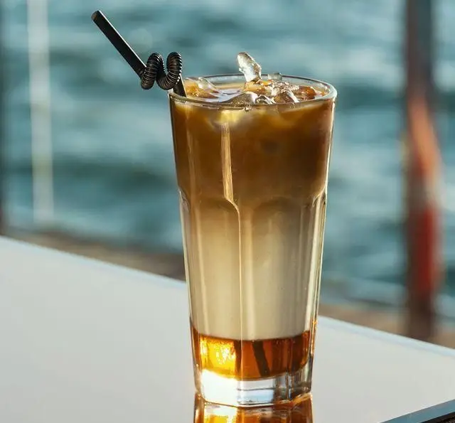 نوشیدنی لاته سرد دلچسب با یخ و سیروپ کارامل در لیوانی بلند، در محیط دلنشین کشتی رستوران رویال لانژ با منظره زیبای دریا.