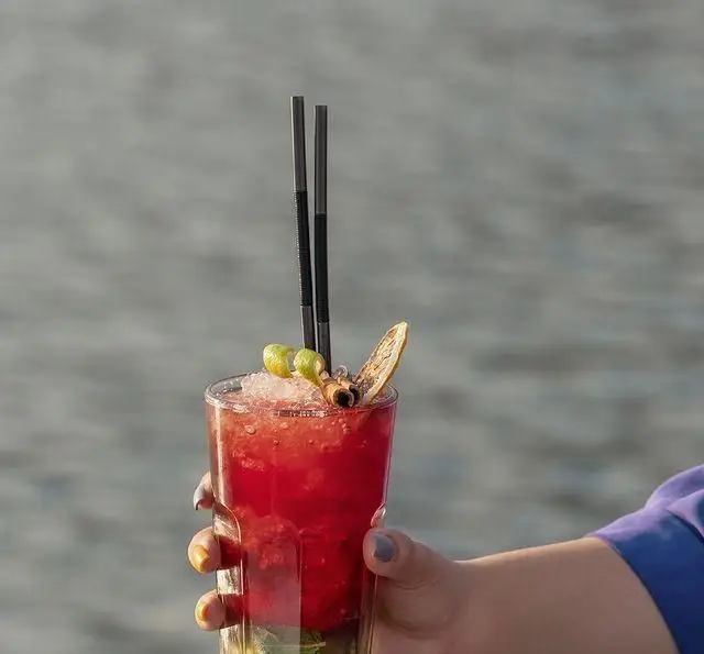 کوکتل خنک و قرمز رنگ کشتی رستوران رویال لانژ، با نی و تزیینات، در دست مشتری و پس‌زمینه منظره آب دریا.