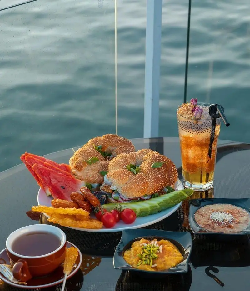 صبحانه مجلل و خوشمزه در کشتی رستوران رویال لانژ تهران، شامل نان کنجدی، میوه، چای، نوشیدنی سنتی و دسرهای ایرانی با منظره دریا