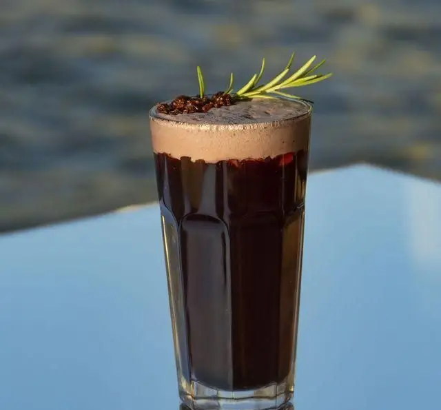 نوشیدنی تیره رنگ خنک با فوم و تزیین رزماری در لیوانی بلند، سرو شده در کشتی رستوران رویال لانژ با دید دریا و فضایی دلنشین.