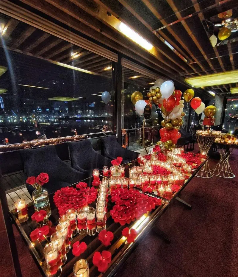 دکوراسیون رمانتیک و لوکس کشتی رستوران رویال لانژ با شمع، گلبرگ قرمز و بادکنک‌ها برای جشن سالگرد و ولنتاین با منظره شب شهر.