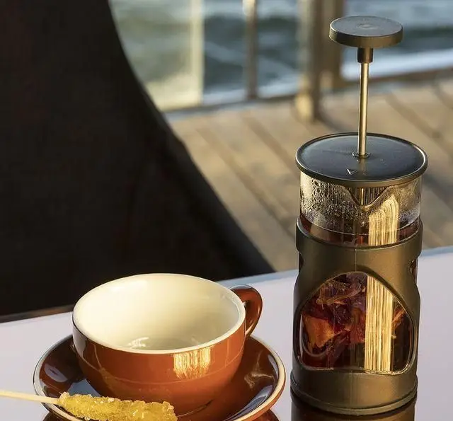 لذت نوشیدن چای یا دمنوش گرم در دمنوش ساز فرنچ پرس در کشتی رستوران رویال لانژ، همراه با فنجان خالی و شکر نبات، با منظره دلنشین آبی.