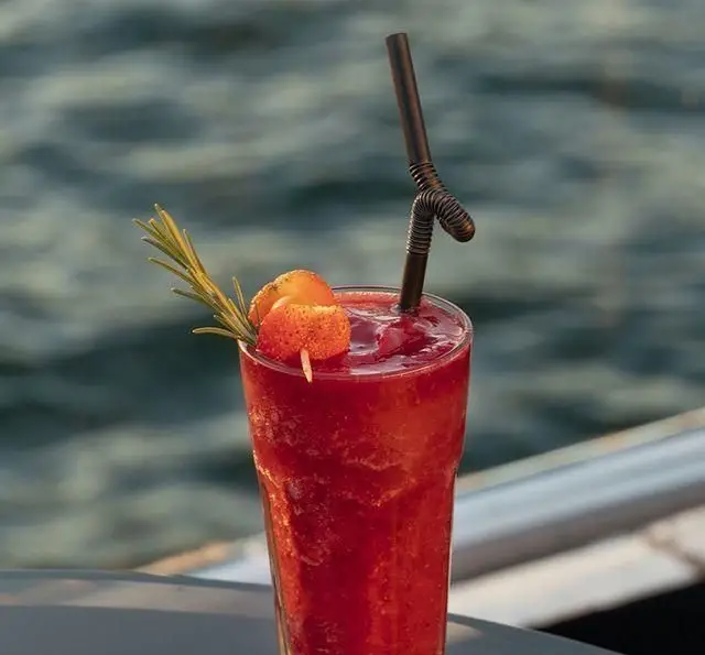 یک لیوان نوشیدنی اسموتی یا کوکتل توت فرنگی قرمز با تزئین رزماری و میوه در کنار آب، نمادی از تجربه دلنشین در کشتی رستوران رویال لانژ.