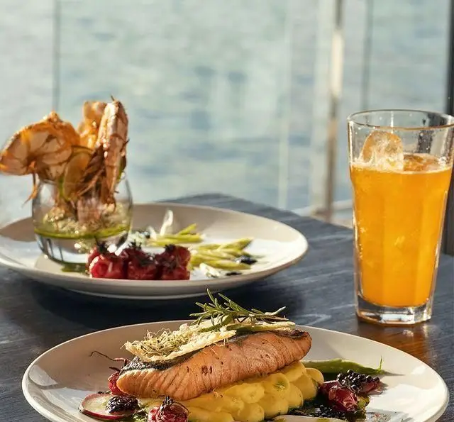 تصویری از غذاهای لذیذ کشتی رستوران رویال لانژ: ماهی سالمون کبابی، میگوی ویژه و نوشیدنی خنک با منظره آبی دلنشین.