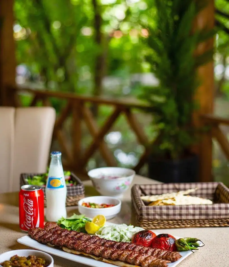 کباب کوبیده معروف کبابسرای شب زدگان با مخلفات کامل، گوجه کبابی، دوغ و نوشابه در فضای سبز و زیبای رستوران.