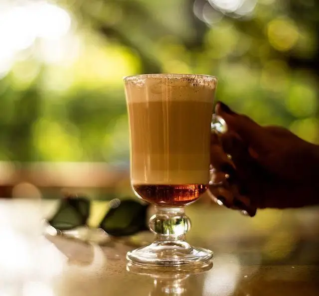 نوشیدنی لایه‌ای دلچسب در لیوان پایه دار، در فضای باز کبابسرای شب زدگان با پس‌زمینه سبز و دلنشین.