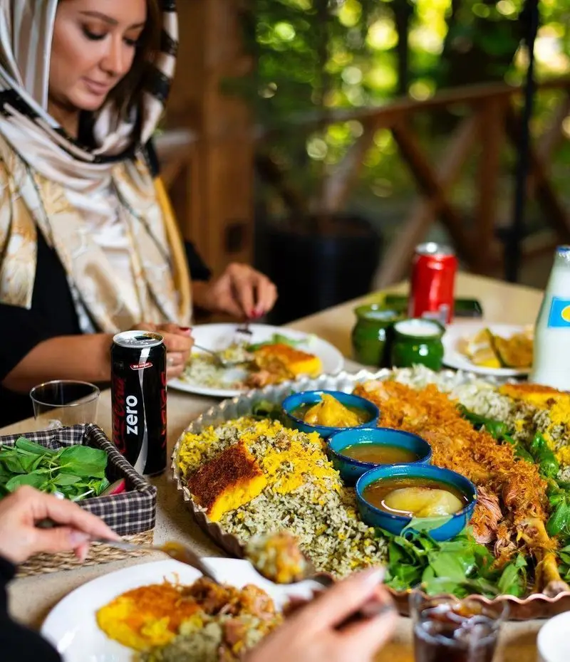 مشتریان در کبابسرای شب زدگان از یک سفره غذای لذیذ ایرانی شامل باقالی پلو، ته دیگ و خورش در فضایی دلپذیر لذت می برند.