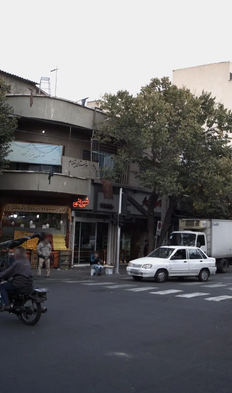 تصویری از نمای بیرونی شیرینی آنالار در خیابان ولیعصر، با نور نئون قرمز و جنب و جوش روزمره شهر، شامل خودروها و درختان.