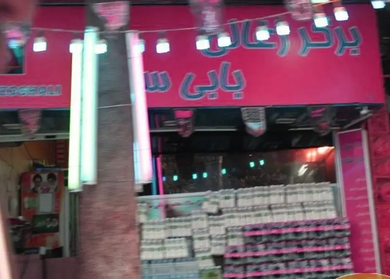 تصویری از نمای بیرونی فست فود بابی ساندز با تابلوی روشن "برگر زغالی و بابی ساندز" و بطری‌های نوشیدنی داخل مغازه.