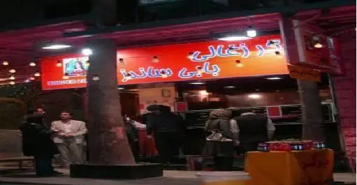 نمای شبانه از مغازه فست فود بابی ساندز با تابلوی روشن «برگر زغالی بابی ساندز» و مشتریان در حال خرید ساندویچ و برگر.