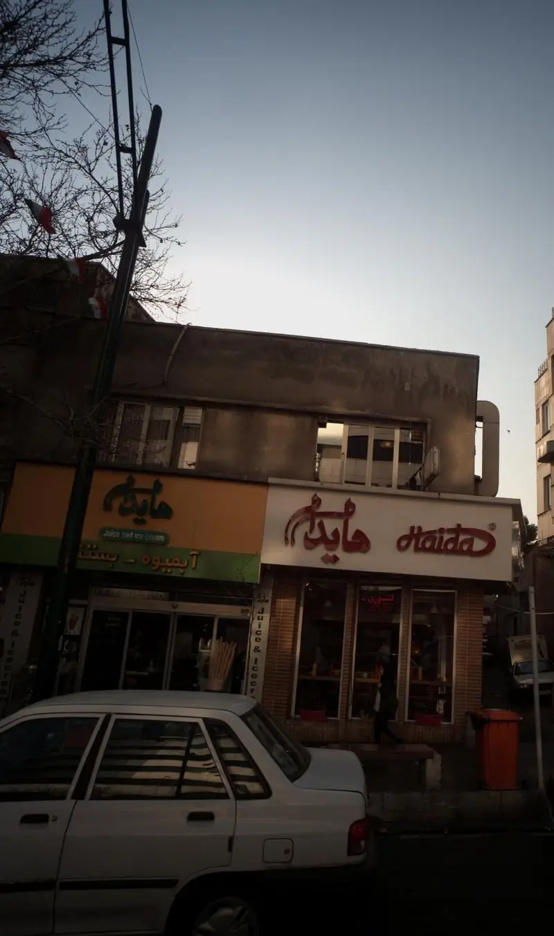 نمای خیابانی از فروشگاه آبمیوه بستنی هایدا با تابلوهای فارسی و انگلیسی، خودروی پارک شده و ساختمان‌های شهری در روز.