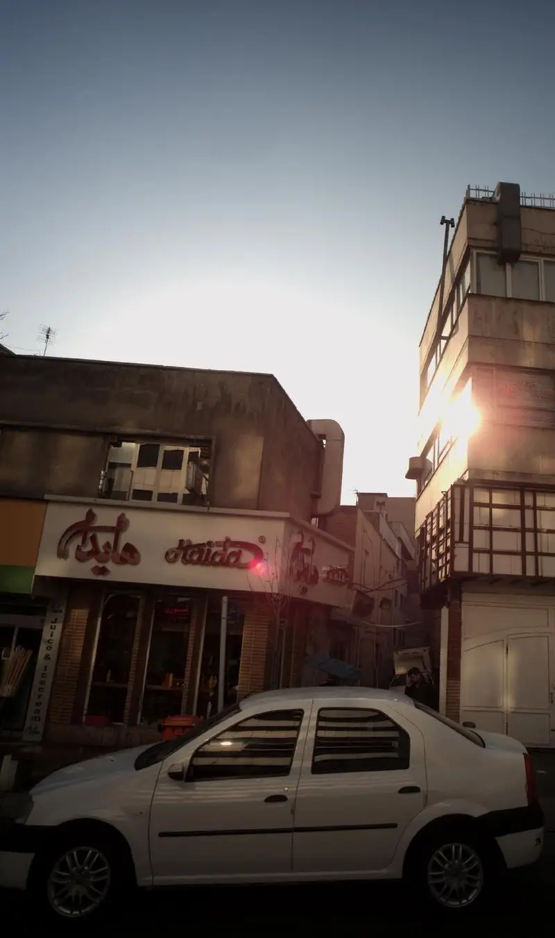 تصویر بیرونی مغازه آبمیوه و بستنی هایدا در خیابان. تابلوی "Haida" با نوشته فارسی و انگلیسی مشخص است. نور خورشید و یک خودروی سفید پارک شده.
