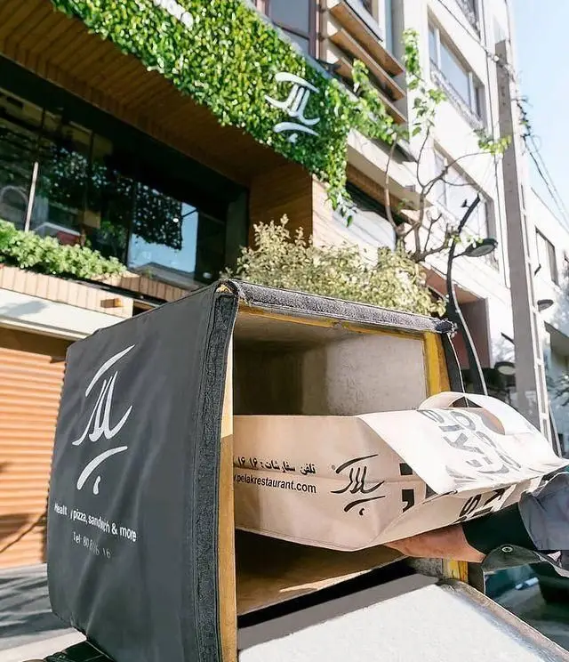 پیک رستوران پلاک در حال قرار دادن بسته غذای سالم، پیتزا و ساندویچ در باکس دلیوری برای ارسال سریع سفارشات مشتریان (pelakrestaurant.com)