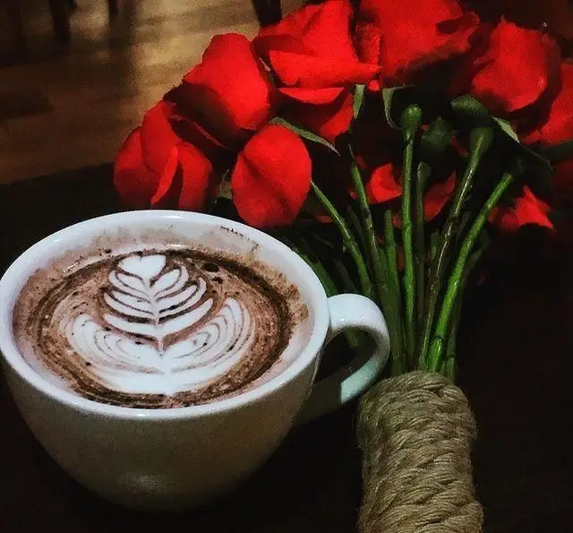 فنجان قهوه لاته با لاته آرت جذاب و دسته گل رز قرمز در کافه رژ، مکانی ایدهآل برای قرارهای عاشقانه و تجربه نوشیدنیهای بینظیر.