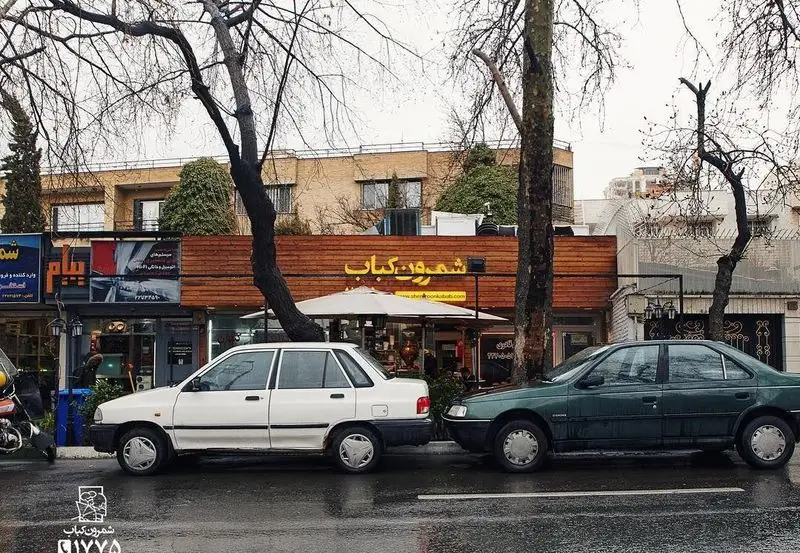 نمای بیرونی و سردر رستوران شمرون کباب در خیابانی پردرخت در تهران. دو خودرو مقابل این کبابی در هوایی بارانی پارک شده‌اند.