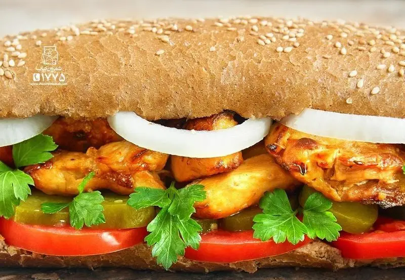 ساندویچ خوش‌طعم و تازه از شمرون کباب: فیله مرغ گریل شده، پیاز، خیارشور، گوجه فرنگی و جعفری روی نان کنجدی قهوه‌ای.
