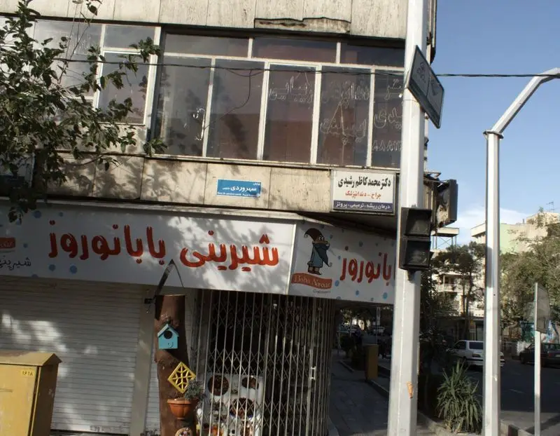 نمای بیرونی قنادی و شیرینی‌فروشی بابا نوروز در خیابان سهروردی تهران، با تابلوی قرمز رنگ و کرکره پایین.