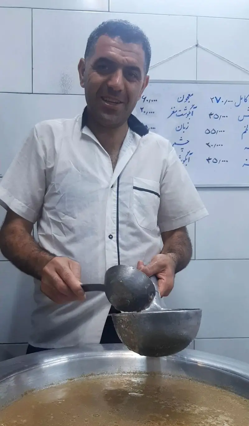 آشپز طباخی پاتوق در حال سرو کله پاچه و آبگوشت سنتی ایرانی از دیگ بزرگ. منوی طباخی با لیست غذاها و قیمت‌ها روی تخته سفید پشت سر او مشخص است.