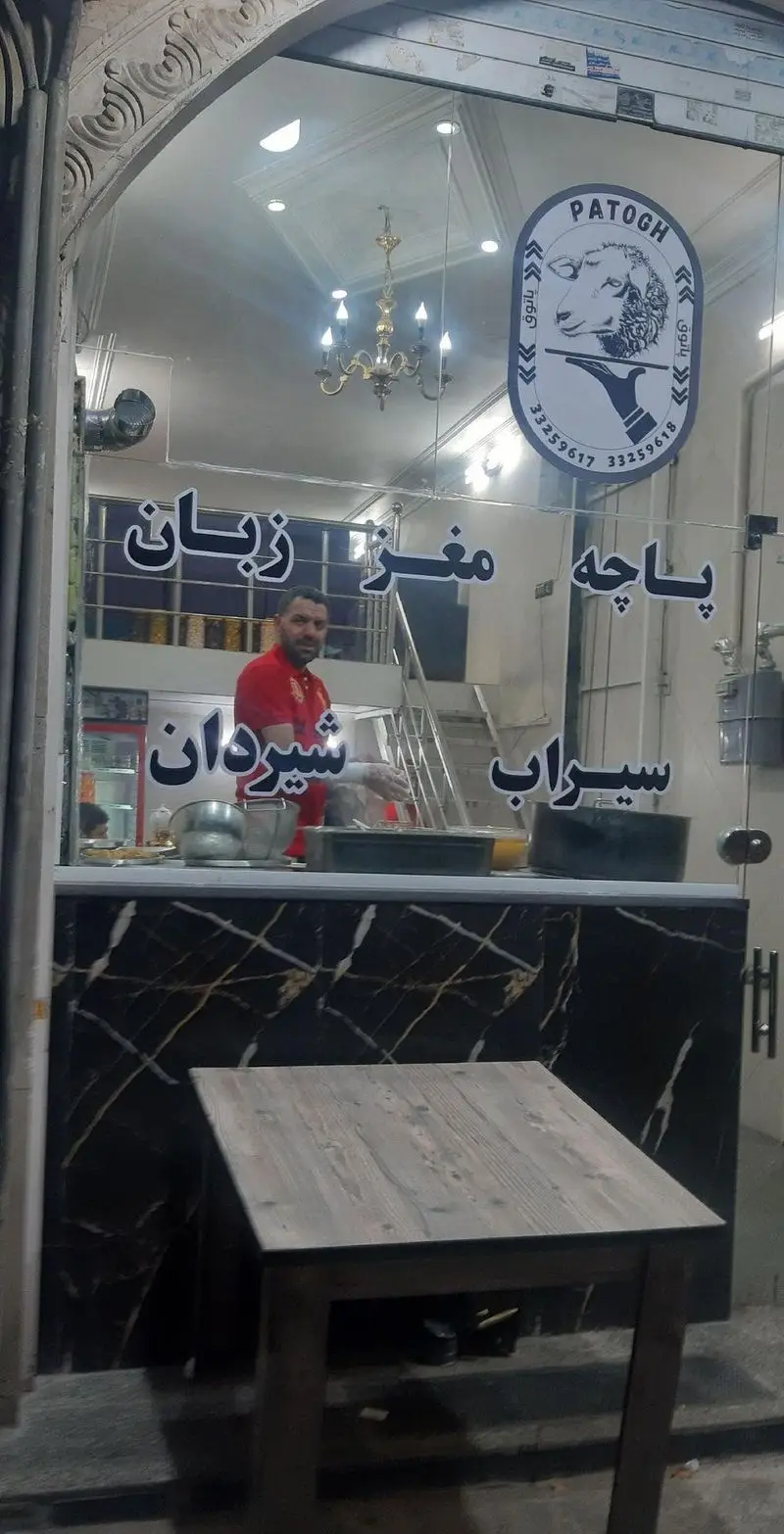 تصویر نمای بیرونی طباخی پاتوق با مردی در حال سرویس‌دهی، میز مشتری و منوی پاچه، مغز، زبان، سیراب و شیردان روی شیشه مغازه.