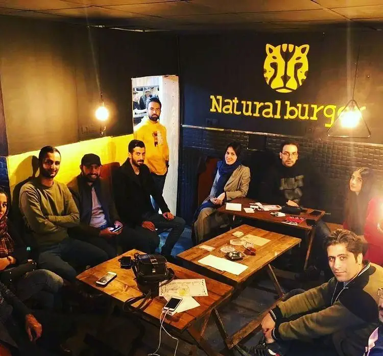 محیط داخلی دلنشین و دوستانه فست فود نچرال برگر با مشتریان نشسته دور میز، لوگوی برجسته و نورپردازی گرم.