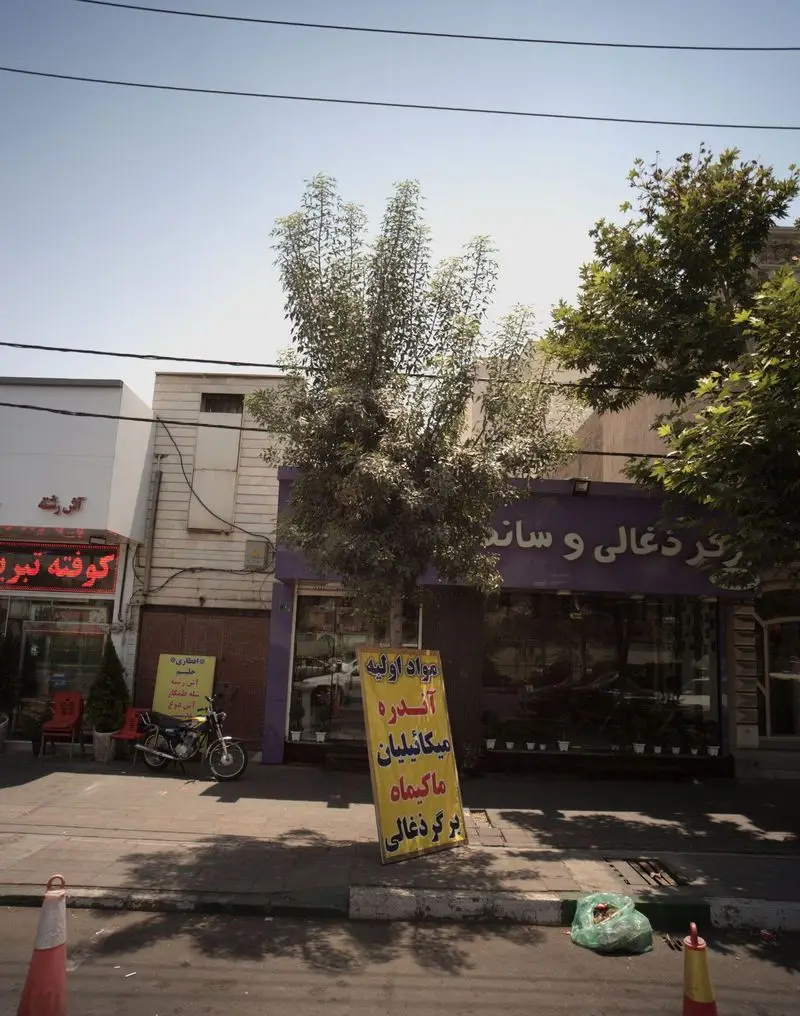 تصویری از نمای بیرونی ممل برگر در یک روز آفتابی، با تابلوی "برگر ذغالی" و مغازه‌های اطراف، همراه با موتورسیکلت و درختان.