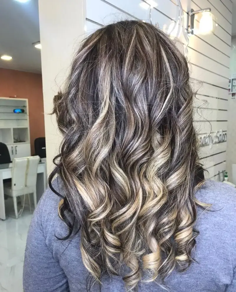 جدیدترین مدل هایلایت موی تیره با رگه‌های طلایی و کرم به صورت فر درشت در سالن زیبایی، مناسب برای ایده رنگ موی دخترانه شیک
