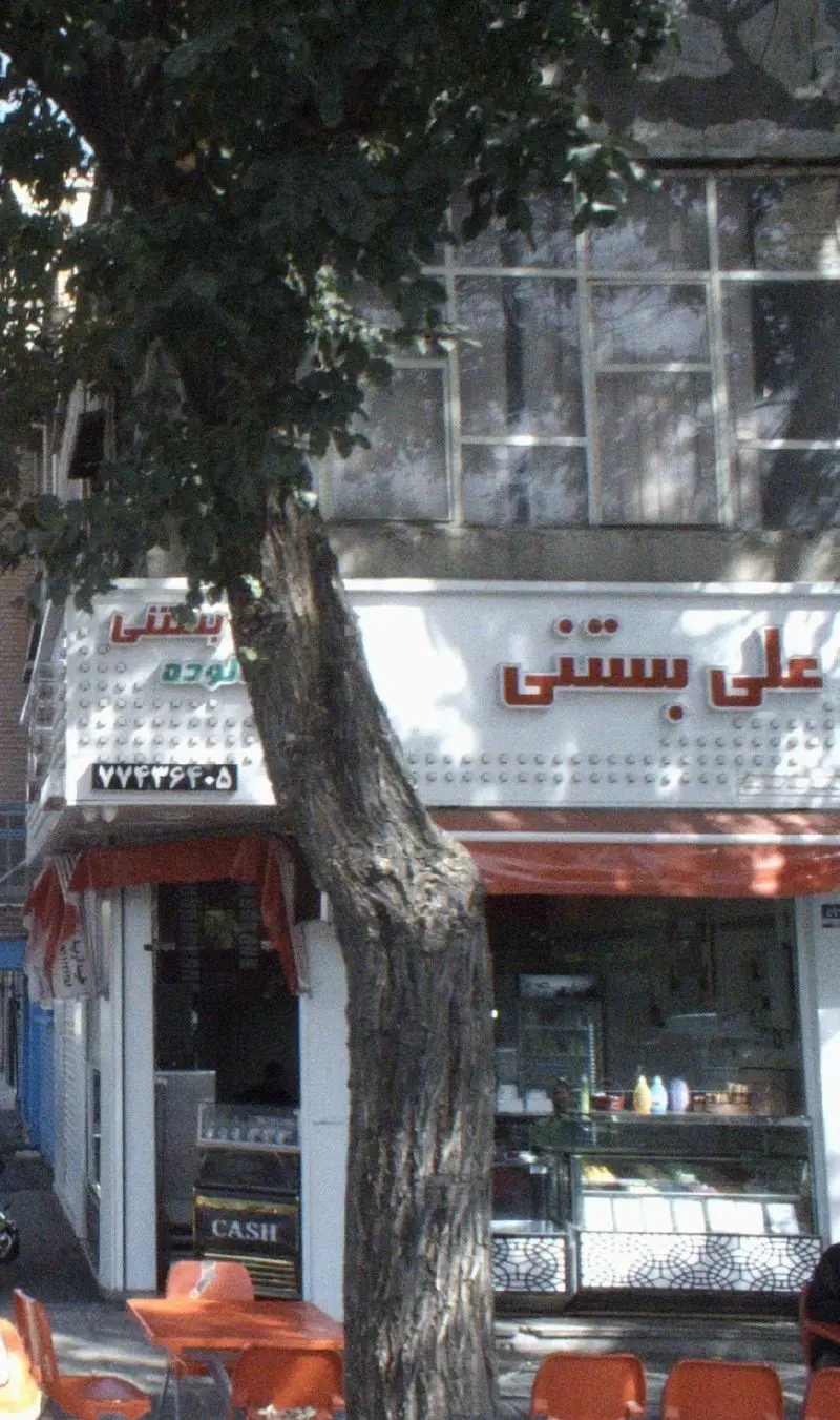 نمای کامل بیرونی مغازه علی بستنی، یک آبمیوه و بستنی فروشی با تابلوی درخشان، درخت سبز در مرکز و میز و صندلی‌های نارنجی در پیاده‌رو.