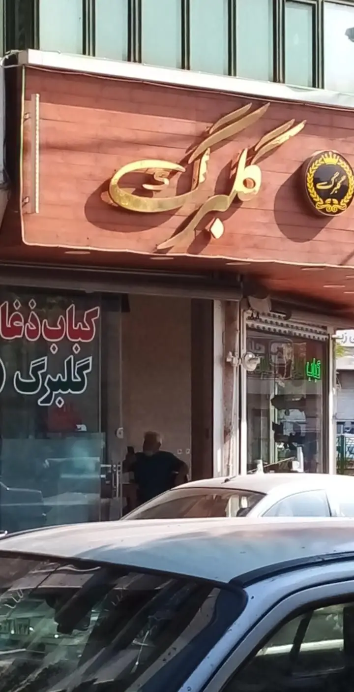 تصویر نمای بیرونی رستوران گلبرگ، کباب ذغالی معروف، با تابلوی شیک و طلایی نام تجاری، ورودی شیشه‌ای و بازتاب نور آفتاب؛ محیطی عالی برای لذت از کباب اصیل ایرانی.