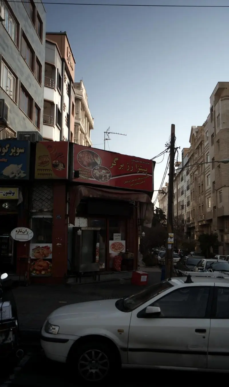 نمای خارجی رستوران پیتزا رز برگر در خیابان، با تابلوی قرمز بزرگ شامل تصاویر غذا، کنار ساختمان‌های شهری و خودروها.