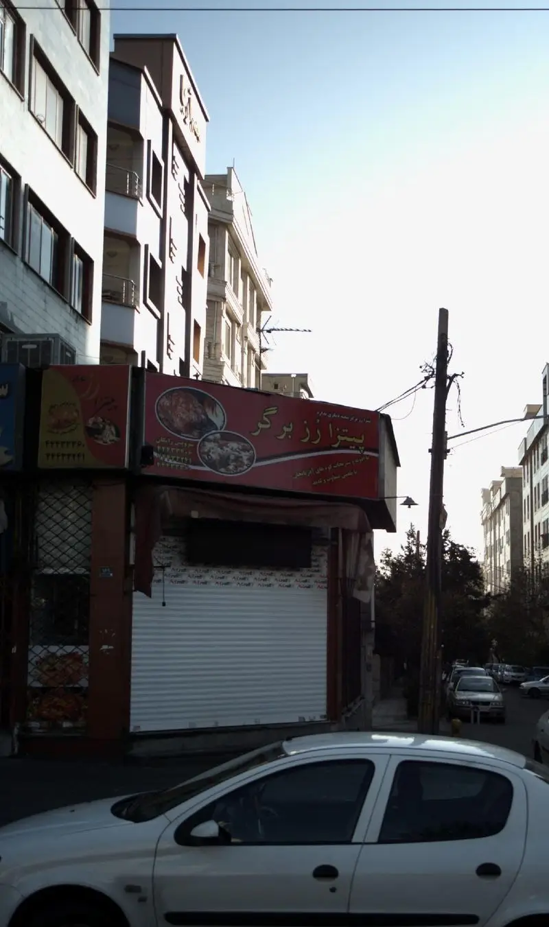 نمای بیرونی فست فود پیتزا رُز برگر در خیابان، با تابلوی قرمز و کرکره بسته، کنار ساختمان‌های بلند و ماشین پارک شده در روز آفتابی.