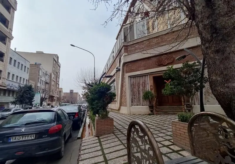 تصویر بیرونی رستوران مسینه با نمای آجری و پنجره‌های بالایی، ورودی چوبی با گلدان و درختان، و ردیفی از ماشین‌ها در حاشیه خیابان.