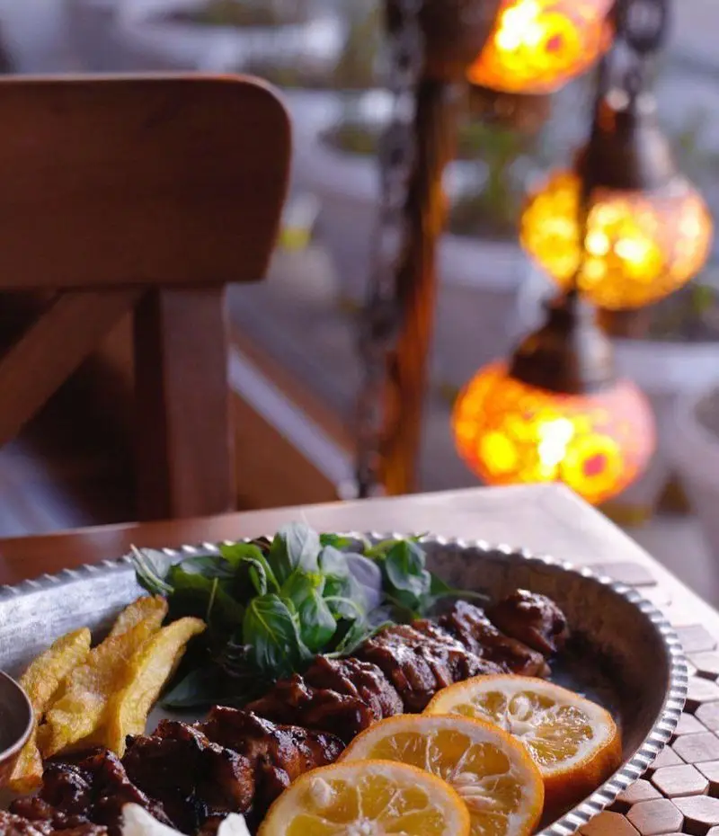 نمای نزدیک از غذای لذیذ رستوران مسینه شامل کباب و سیب‌زمینی با سالاد و پرتقال. دکوراسیون سنتی و فانوس‌های رنگی اتمسفر دلنشینی را ایجاد کرده‌اند.