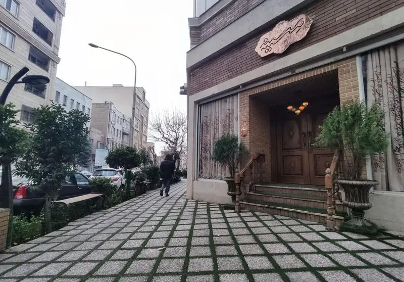 ورودی و نمای بیرونی رستوران مسینه با تابلوی مسی‌رنگ، درب‌های چوبی، پله‌ها، پیاده‌روی سنگی-چمنی و درختان کنار خیابان.