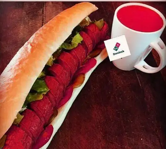 ساندویچ بزرگ سوسیس یا کالباس همراه با کاهو و خیارشور، کنار یک ماگ سفید با نوشیدنی قرمز و برچسب لوگوی دومینوز پیتزا.