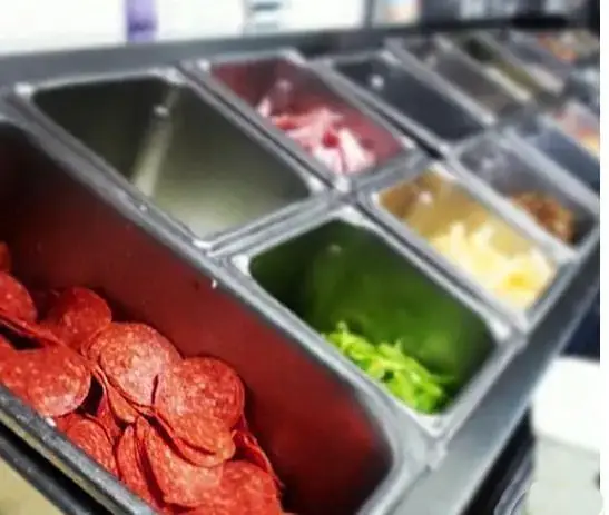 ظرف های استیل حاوی مخلفات متنوع و تازه پیتزا شامل پپرونی، فلفل سبز، و انواع پنیر در خط آماده سازی دومینوز پیتزا.