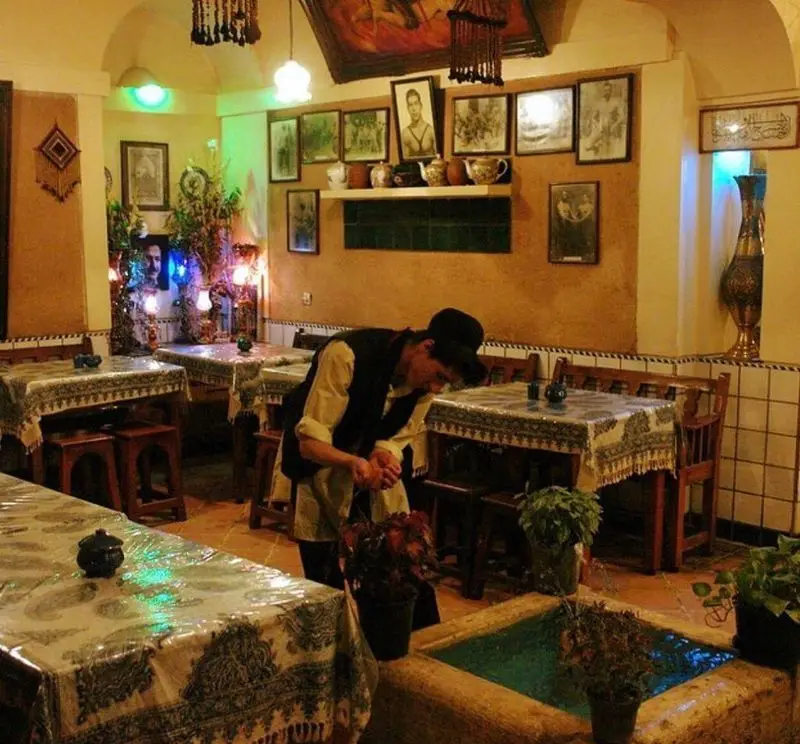 فضای داخلی دنج و سنتی رستوران ایرانی آذری با دکوراسیون اصیل، میزهای چیده شده و مردی در حال مراقبت از گلدان‌ها کنار حوض آب.