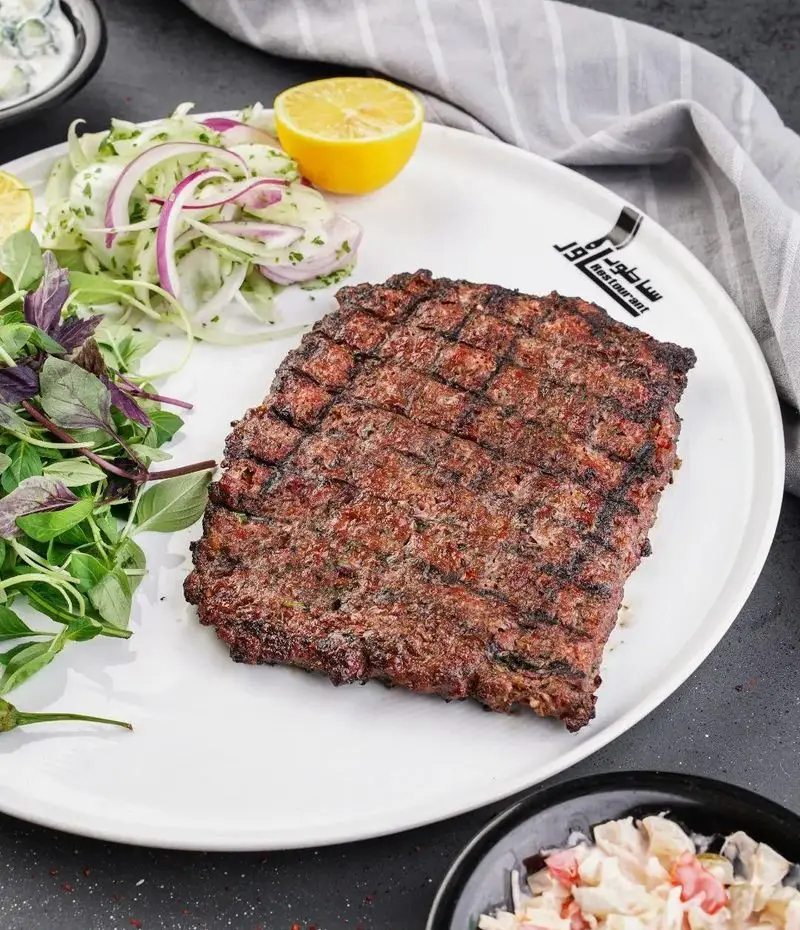 کباب کوبیده گوشت لذیذ رستوران ساطور با رد گریل، همراه سبزی تازه، پیاز، لیمو و سالاد روی بشقاب سفید. تجربه‌ای دلنشین از غذای اصیل ایرانی.