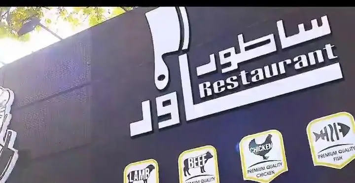 نمای نزدیک از تابلوی رستوران ساطور با لوگوی ساطور و عنوان "Restaurant"، نمایشگر برچسب‌های کیفیت عالی برای بره، گوشت گاو، مرغ و ماهی.