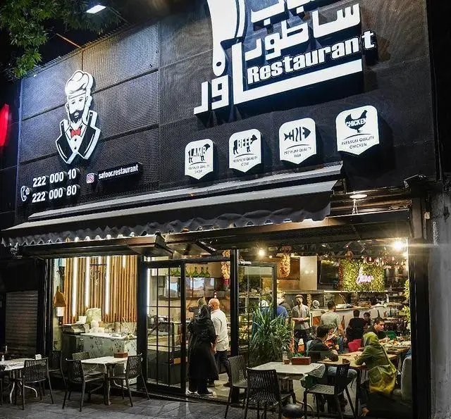 رستوران ساطور: نمای بیرونی شبانه و مدرن با لوگوی سرآشپز، نشانه‌های گوشت، مرغ و ماهی. محیطی دلپذیر برای لذت بردن از غذای باکیفیت.